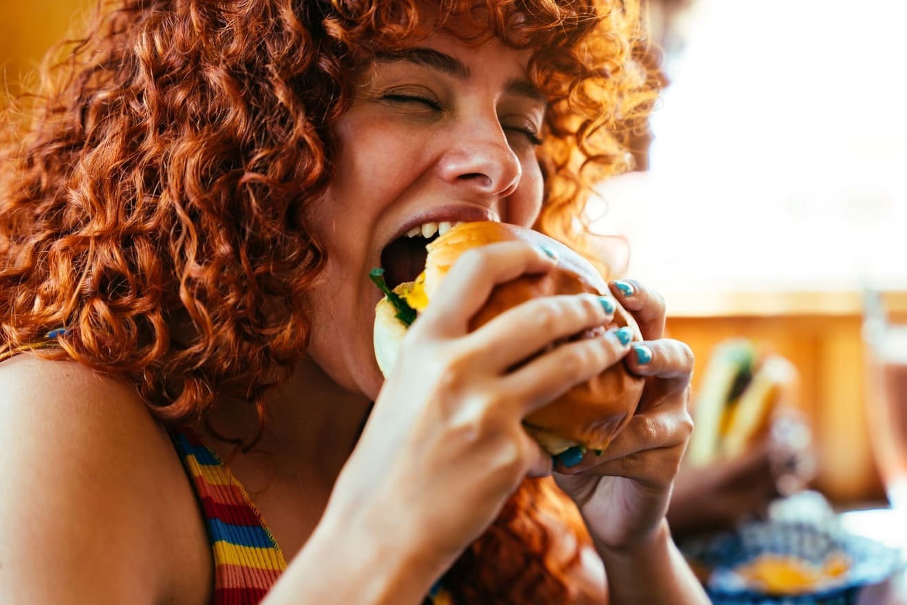 Mujer pelirroja comiendo una hamburguesa grande, representando consumo de comida abundante o alimentos potencialmente pesados para la digestión.