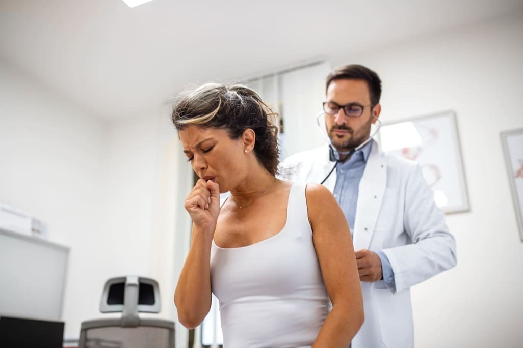 Paciente adulta tosiendo con médico en consulta, mientras el doctor realiza una auscultación a la paciente.