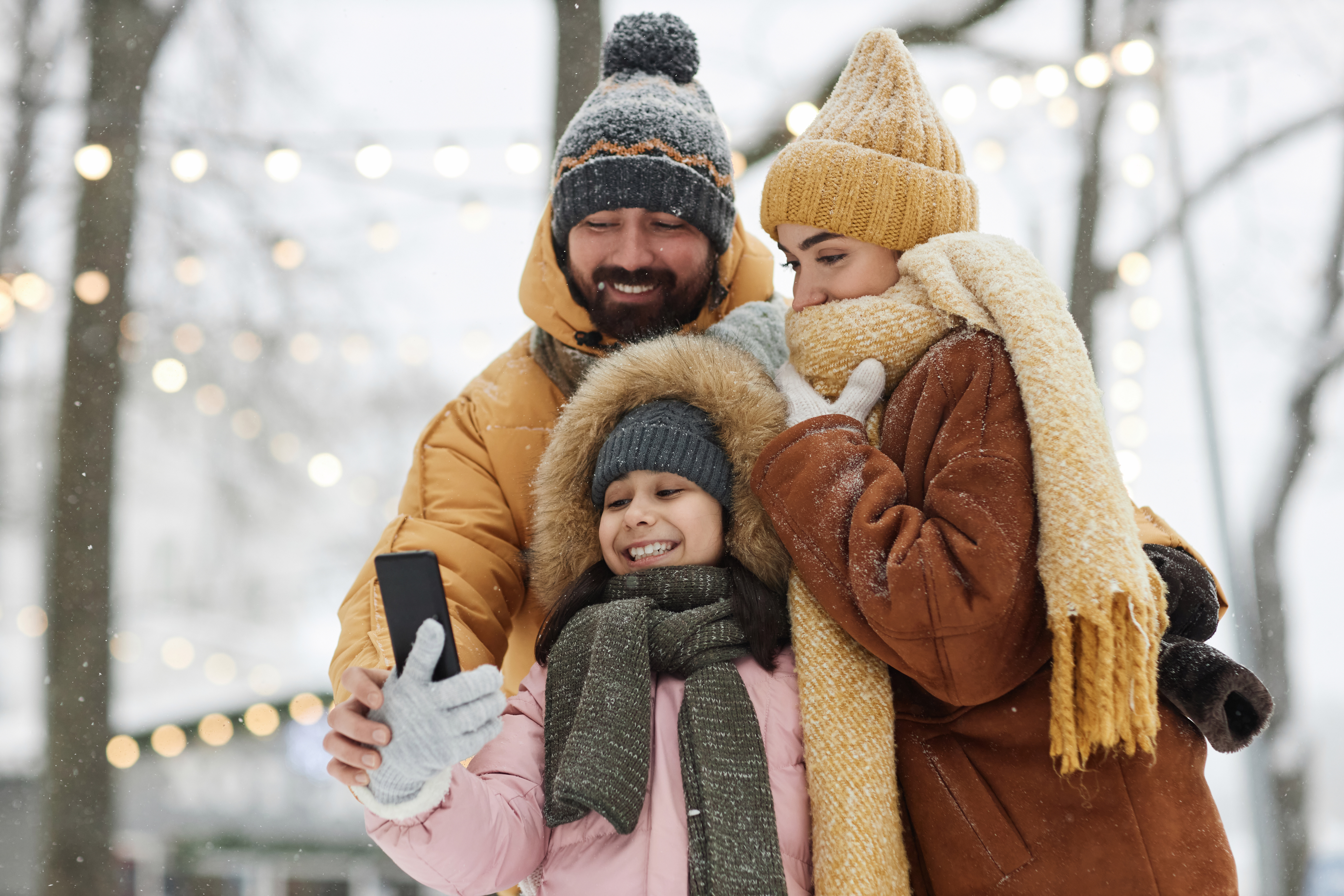 Padres e hija sonríen mientras se toman una selfie juntos en un ambiente nevado y festivo.