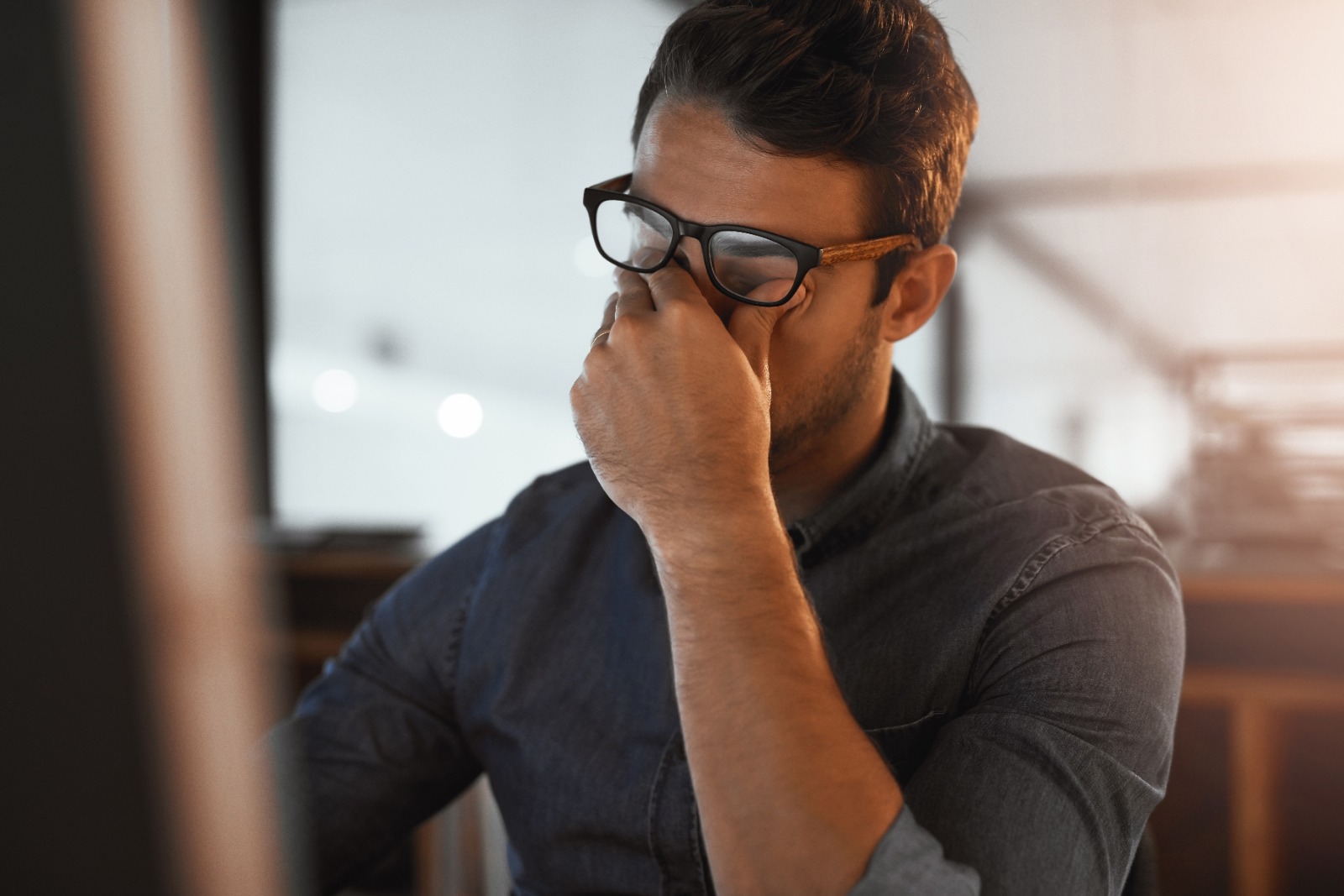 Hombre joven con gafas mostrando signos de ansiedad o estrés mientras trabaja frente a un ordenador, tocándose el rostro con expresión de agotamiento