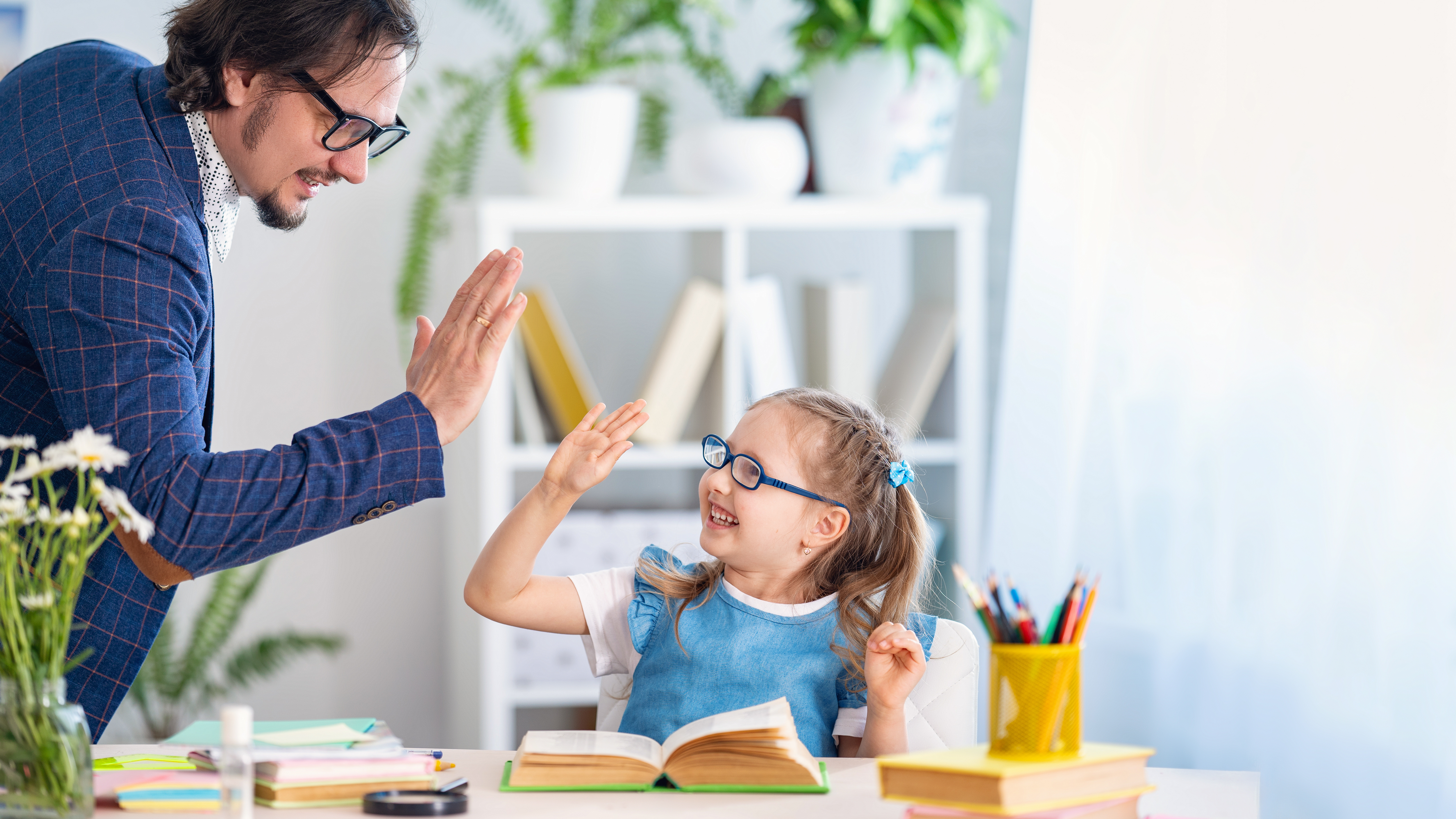 Hombre adulto y niña con gafas se dan un 'choca esos cinco' mientras estudian juntos en un ambiente alegre y educativo.